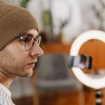 Criador grava um vídeo com smartphone diante de uma luz circular em um ambiente doméstico.