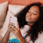 Jovem de cabelo cacheado usa um smartphone enquanto exibe unhas pintadas com cores variadas.