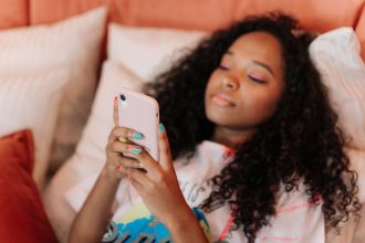Jovem de cabelo cacheado usa um smartphone enquanto exibe unhas pintadas com cores variadas.