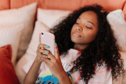 Jovem de cabelo cacheado usa um smartphone enquanto exibe unhas pintadas com cores variadas.